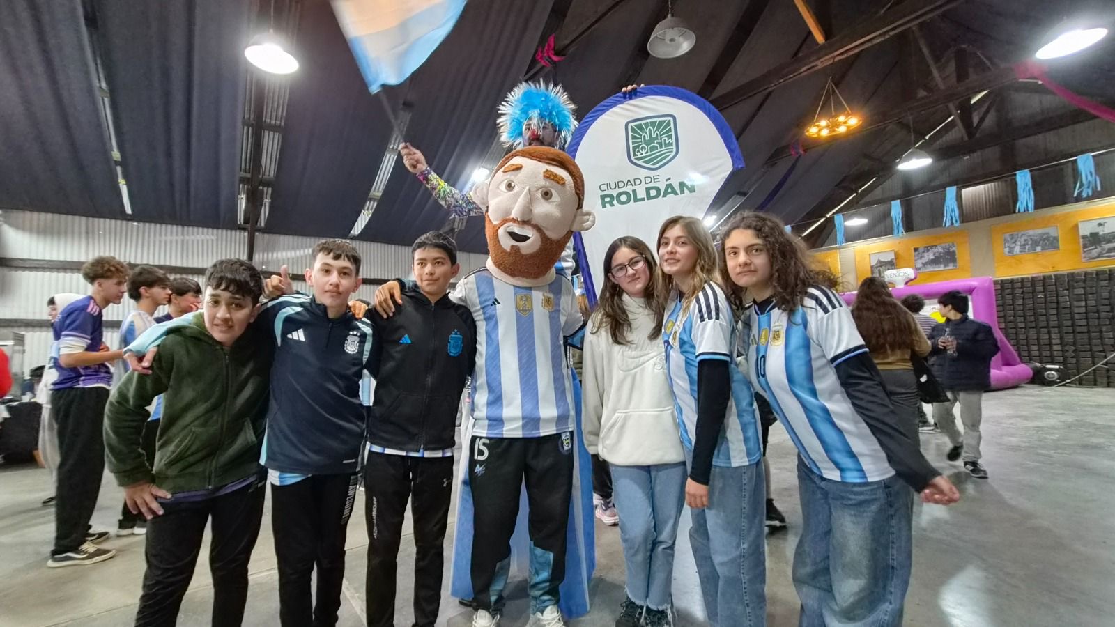 Jóvenes posan con la mascota de Messi en el Fan Fest de Roldán junto al cartel de la Ciudad de Roldán