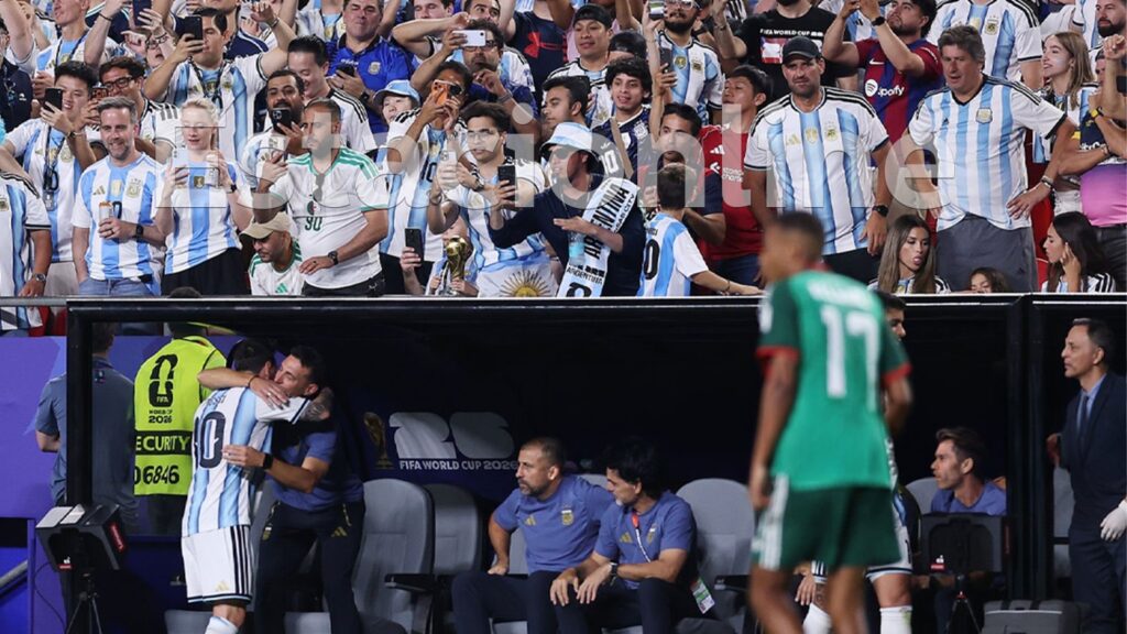 Roly Santacroce con la camiseta argentina en la tribuna, detrás del abrazo entre Messi y Scaloni en el Mundial 2026