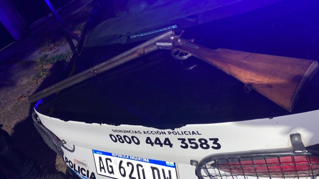 Una escopeta de doble caño secuestrada por la Policía, apoyada sobre el capot de un móvil policial