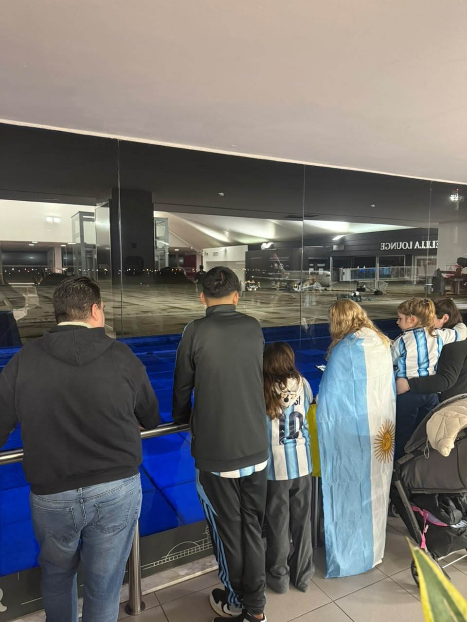 Una familia con la bandera argentina y camisetas de la Selección mira la pista desde el ventanal del aeropuerto