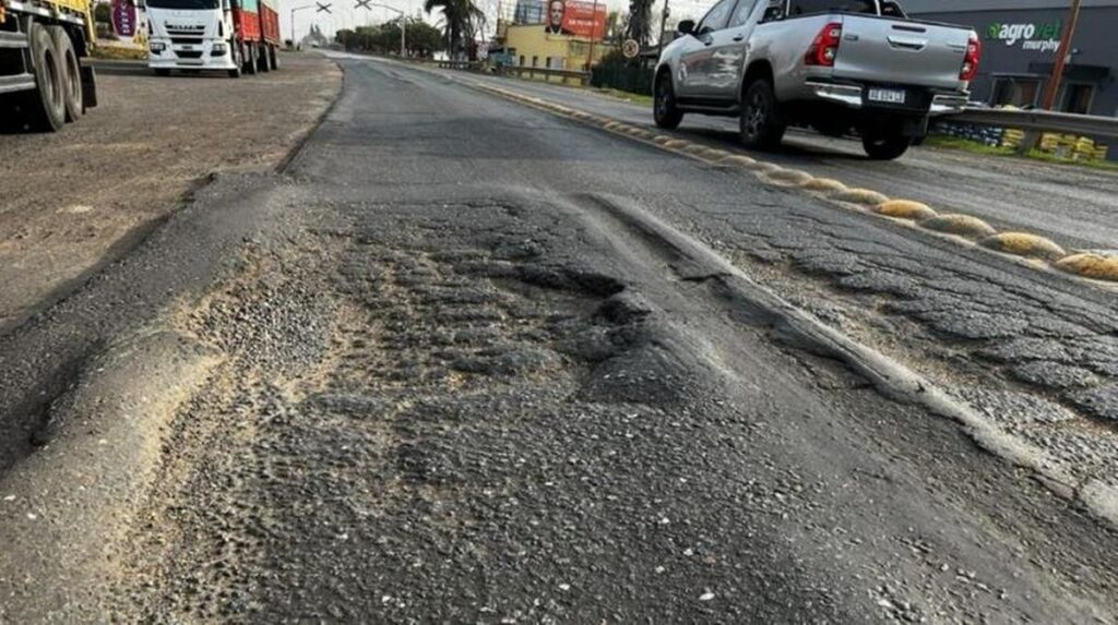 La calzada de la ruta A012 con baches, hundimientos y asfalto deformado, con camiones circulando al costado