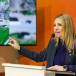 Coudannes: “Santa Fe avanza en paritarias y recomposición salarial semestre a semestre” Virginia Coudannes habla en un atril frente a una pantalla con imágenes satelitales de una ruta